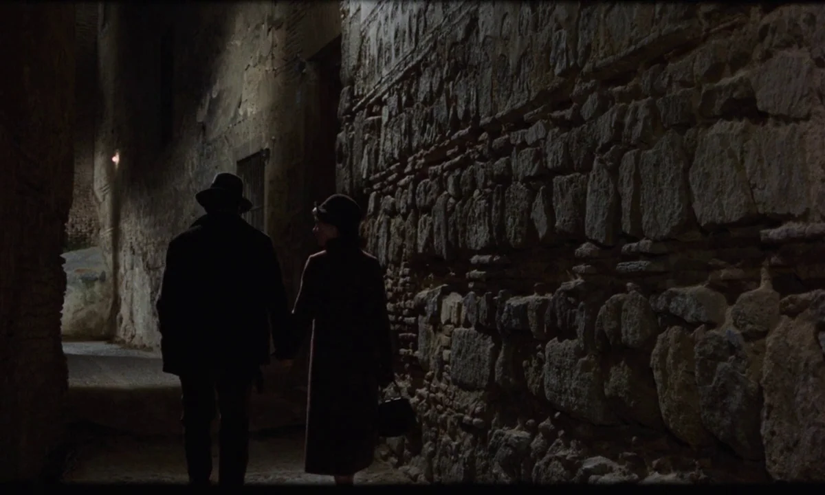 Tristana (1970) movie still: fullbody — A narrow, dark stone alleyway features uneven walls built from stacked rough masonry. A man and a woman in…