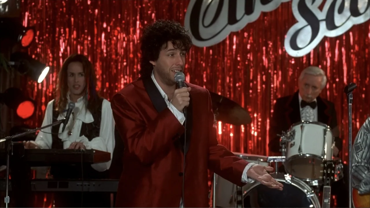 The Wedding Singer (1998) movie still: medium — A man stands on a stage in front of shiny red metallic streamers. The man is singing into a silver…
