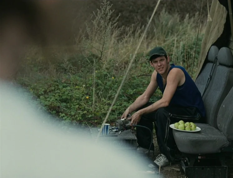 Fish Tank (2009) movie still: medium — An outdoor area features two detached automotive bucket seats placed on a grassy patch. A young man sits on…