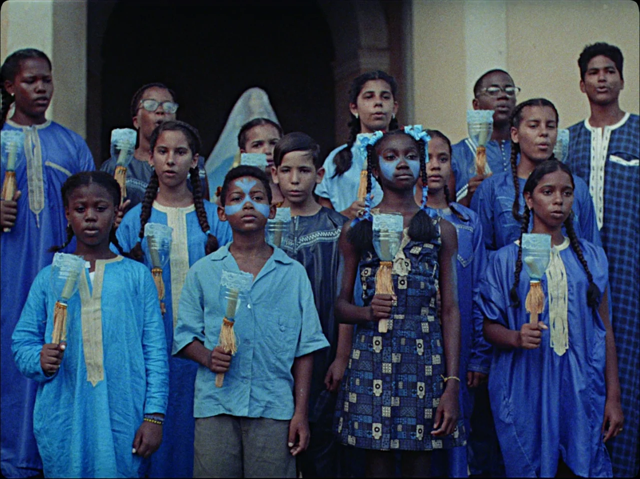 Guava Island (2019) movie still: medium — A group of children and teenagers stands together in an outdoor courtyard area. They wear matching blue…