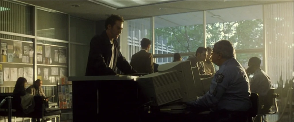Fight Club (1999) movie still: medium — A modern reception area features large windows, a notice board, and beige interior decor. A man stands…