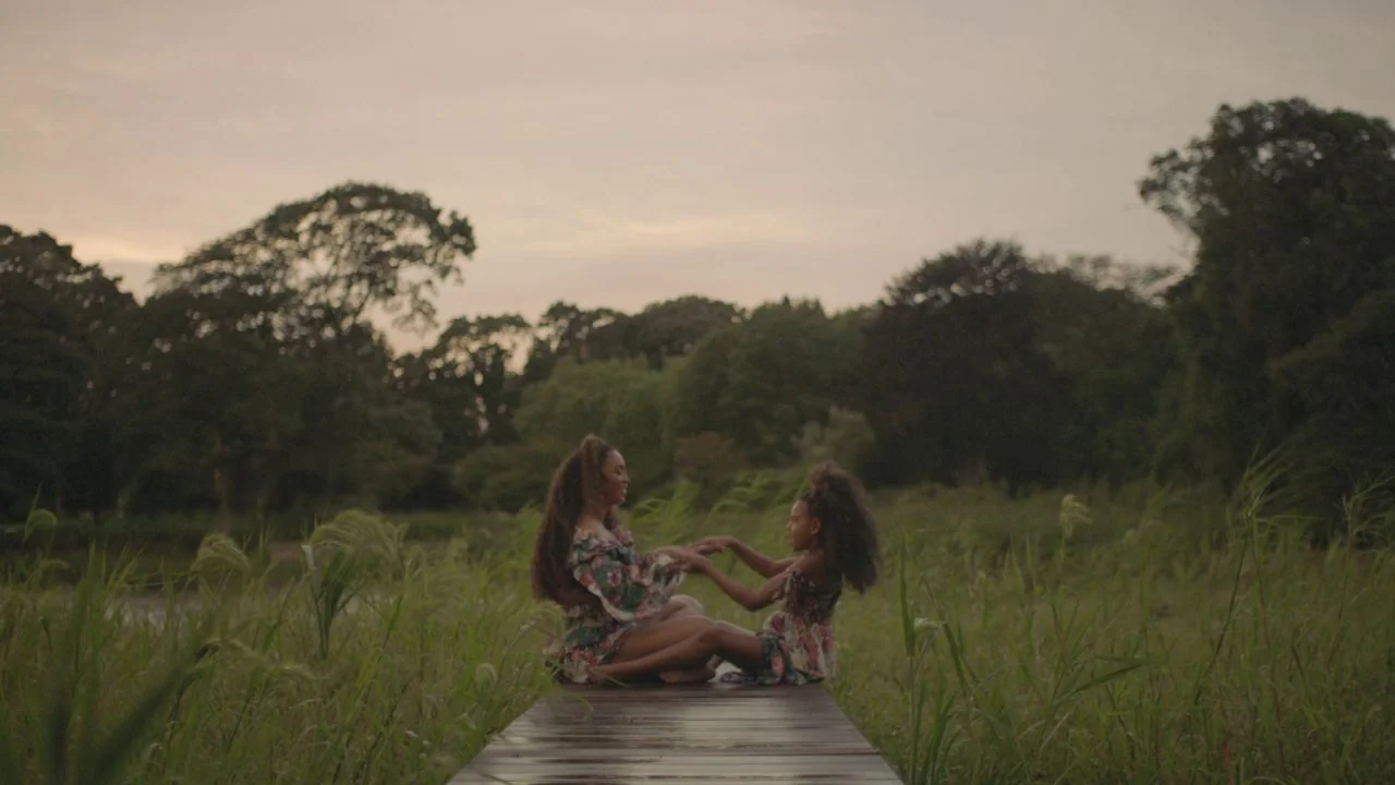 Black is King (2020) movie still: wide — A wooden dock extends into a grassy wetland surrounded by dense, tall trees. A woman and a young girl sit…