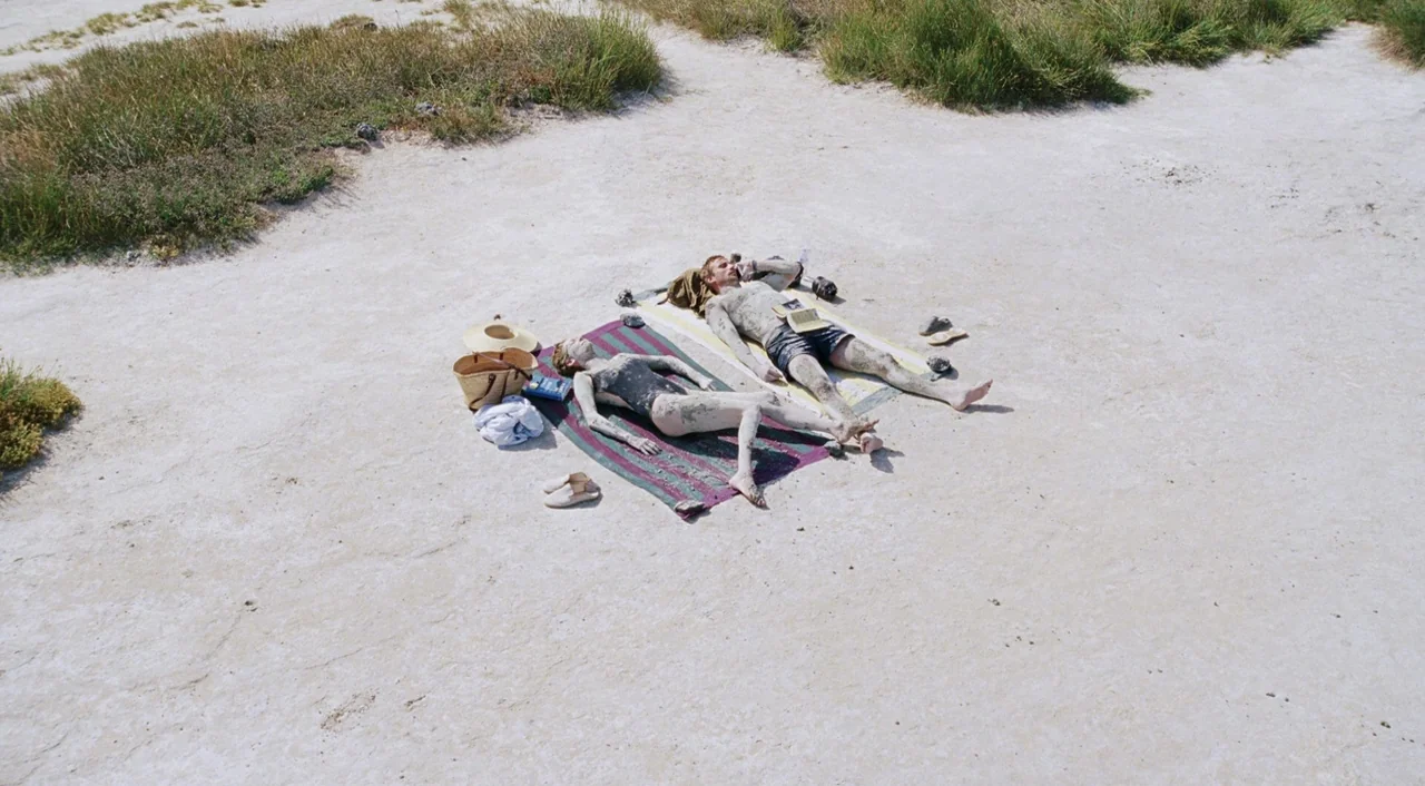 A Bigger Splash (2015) movie still: wide — Two people lie on towels in the middle of a vast white salt flat. Both are coated in gray mud and resting…