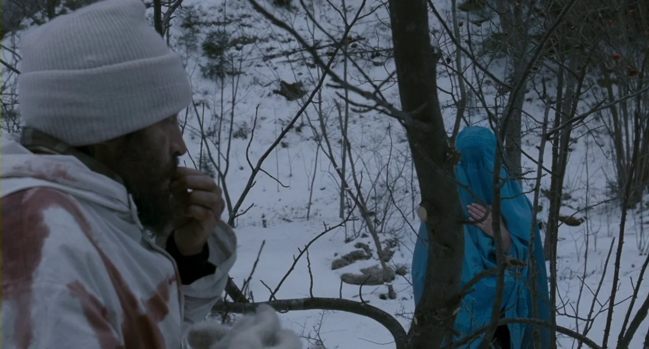 Essential Killing (2010) movie still: medium — A snowy, sparse woodland area with leafless trees and patches of frozen ground. A bearded man in a…
