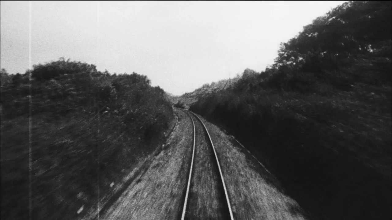 Mysterious Object at Noon (2000) movie still: establishing — A set of parallel railway tracks curves through a hilly, vegetated landscape. The scene…