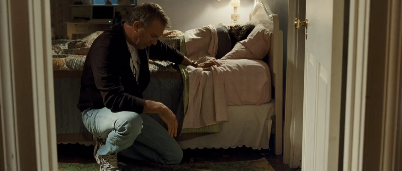 The New Daughter (2009) movie still: medium — A bedroom contains a white wooden bed frame and a patterned quilt. A middle-aged man kneels beside the…