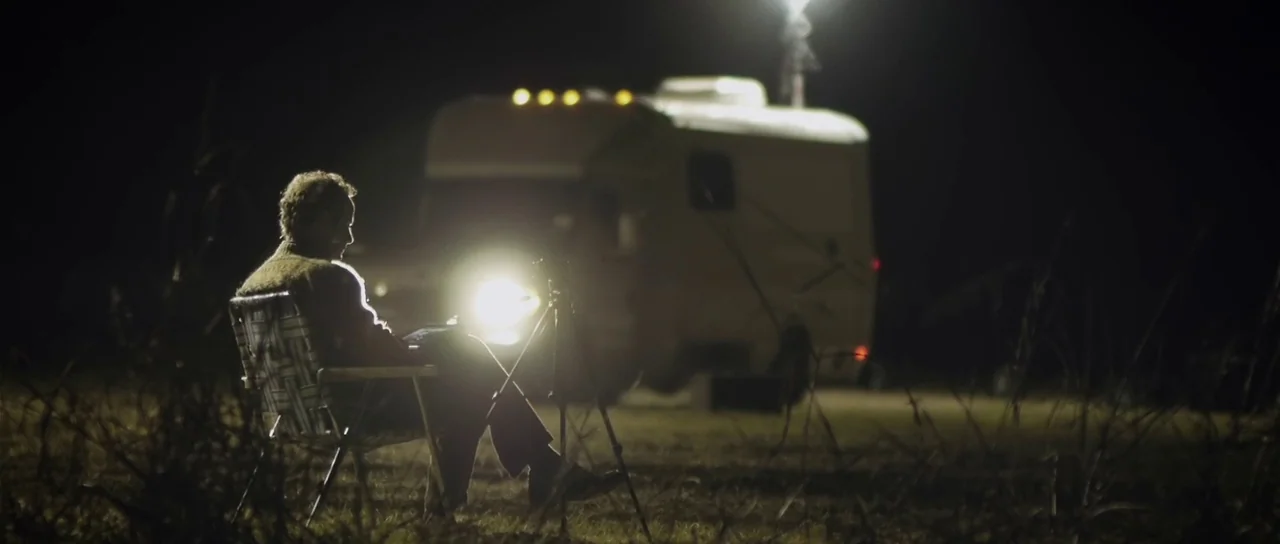 Upstream Color (2013) movie still: wide — A dark, grassy field at night holds a parked camper vehicle. A man sits in a folding chair using a laptop…