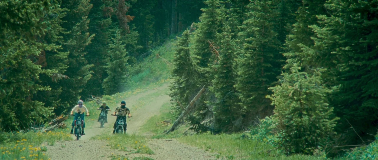 Riddle of Fire (2023) movie still: wide — A winding dirt path traverses through a dense forest of evergreen pine trees. Three individuals ride small…