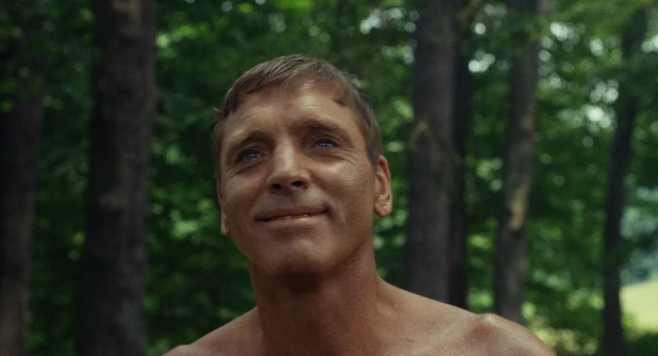 The Swimmer (1968) movie still: closeup — A dense forest environment with tall, blurred tree trunks in the background. A shirtless man stares…