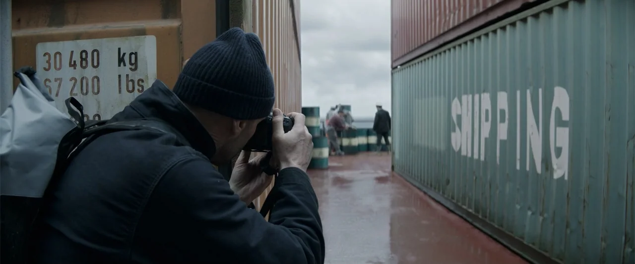 Meg 2: The Trench (2023) movie still: medium — A cargo ship deck features rows of metal shipping containers under a cloudy sky. A man in a dark…