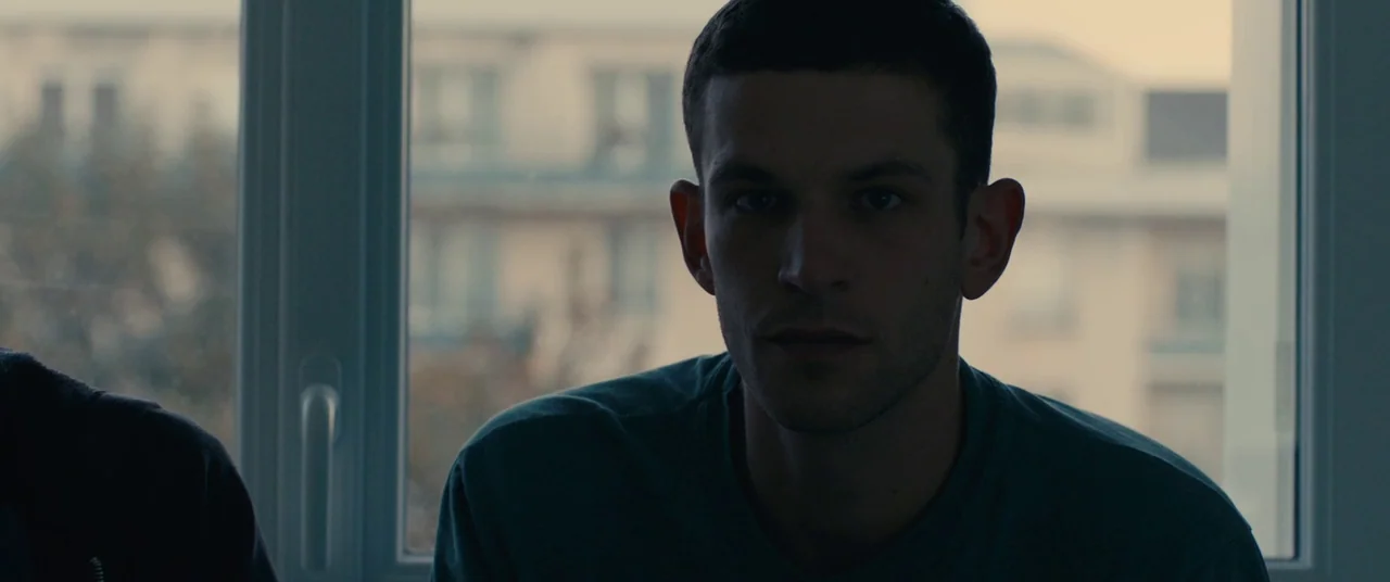 BPM (Beats Per Minute) (2017) movie still: medium — A man sits indoors in front of a white window frame overlooking blurred buildings. He stares…