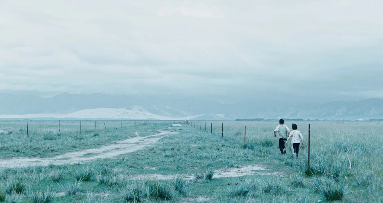 Balloon (2019) movie still: wide — A vast, flat grassland extends toward distant mountains under a heavy overcast sky. Two young children run…