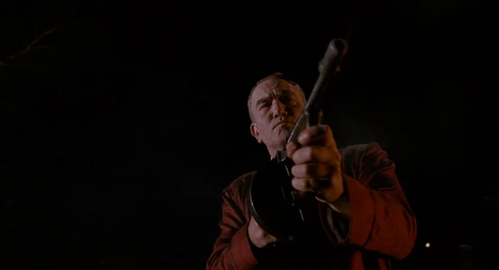 Miller’s Crossing (1990) movie still: medium — A man stands in an undifferentiated darkness holding a firearm pointed toward the viewer. The man…