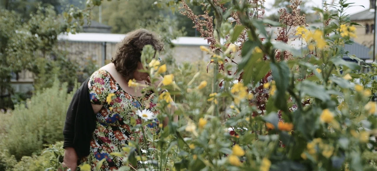 10 Billion: What's On Your Plate (2015) movie still: medium — A woman in a floral dress stands in a lush outdoor plant nursery. She is partially…