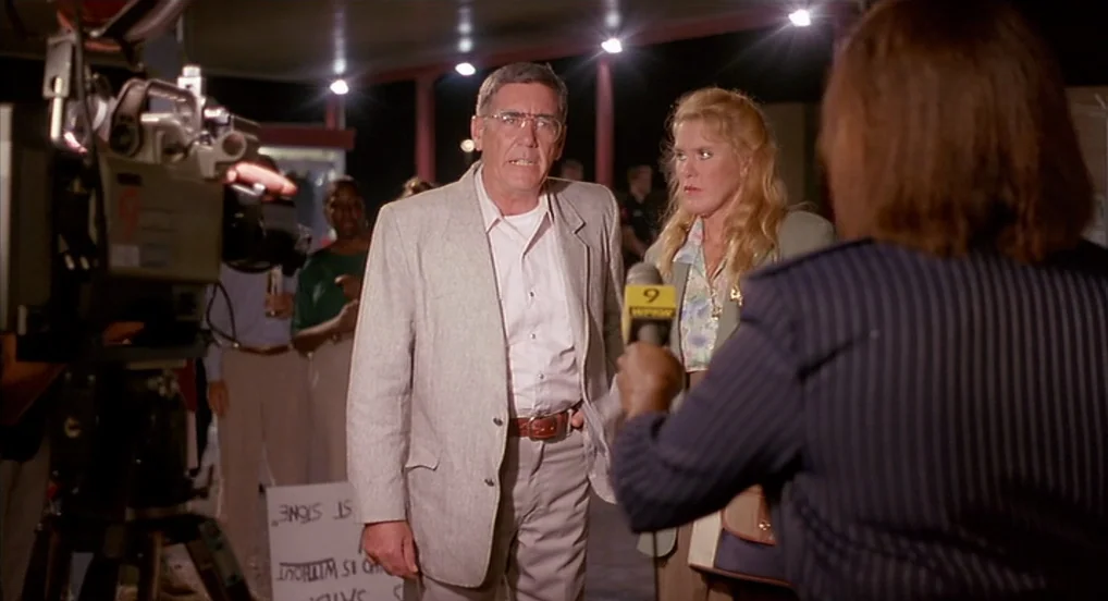 Dead Man Walking (1995) movie still: medium — A dark outdoor parking lot at night with overhead lights illuminating a news interview. A man stands…
