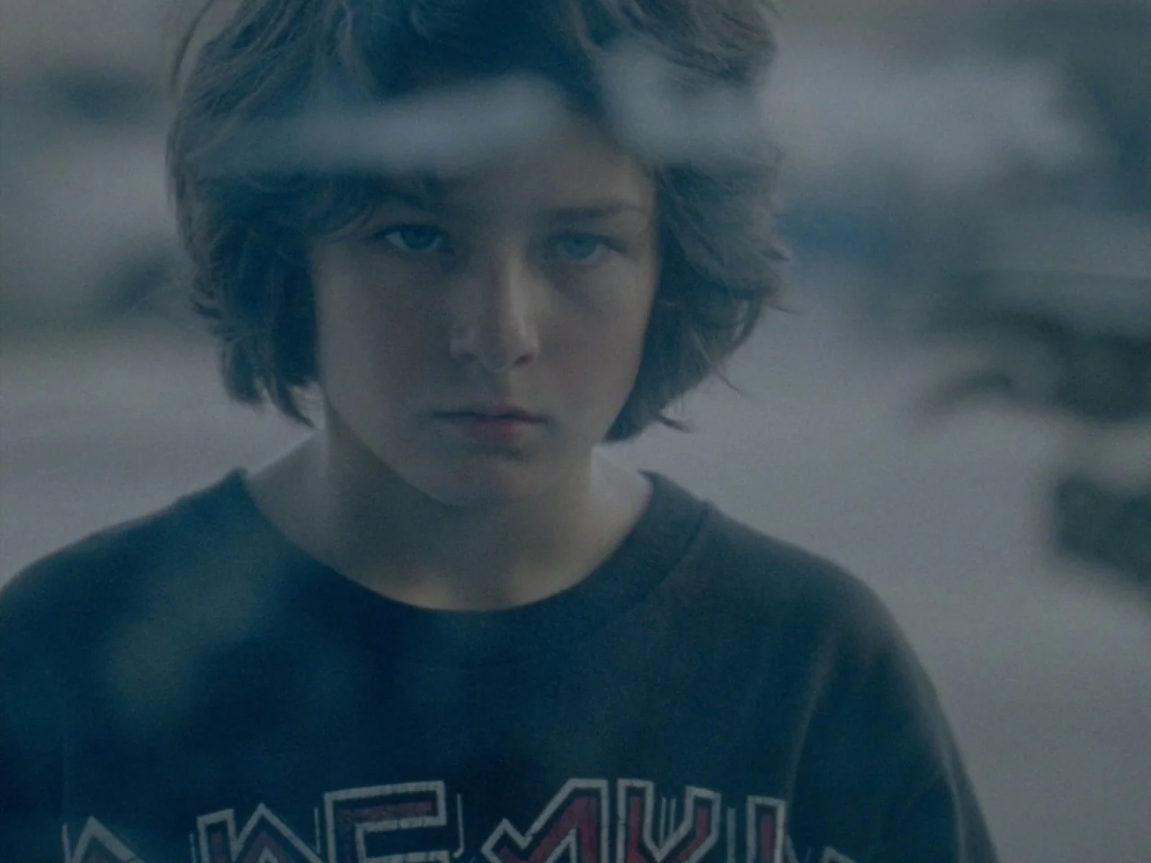 Mid90s (2018) movie still: closeup — The subject stands behind a pane of glass on an urban city street. The boy stares directly forward with a stoic…