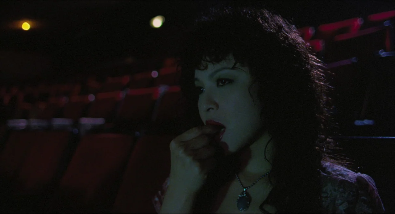 Goodbye, Dragon Inn (2003) movie still: closeup — A dark movie theater interior with rows of out-of-focus red seating in the background. A woman…