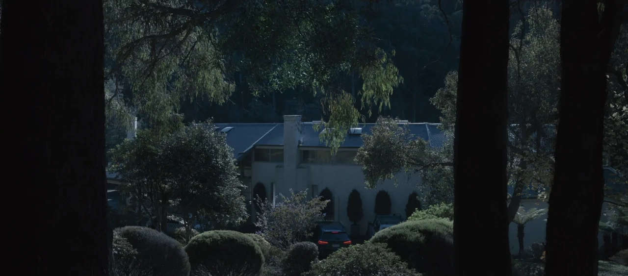 Force of Nature: The Dry 2 (2024) movie still: establishing — A low-rise modern house with a dark roof sits tucked among dense foliage. Two tall…