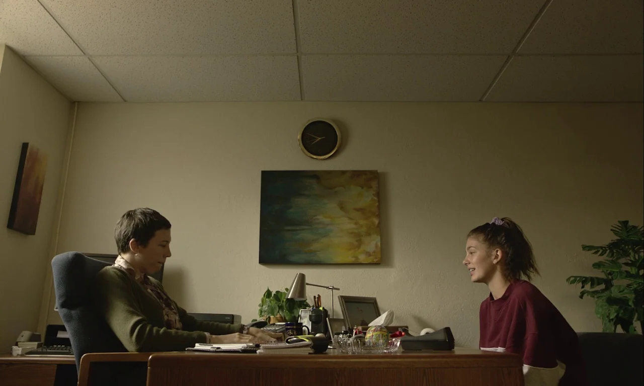 Mickey and the Bear (2019) movie still: medium — An office features beige walls, a wooden desk, and a drop ceiling. A woman sits at the desk while a…