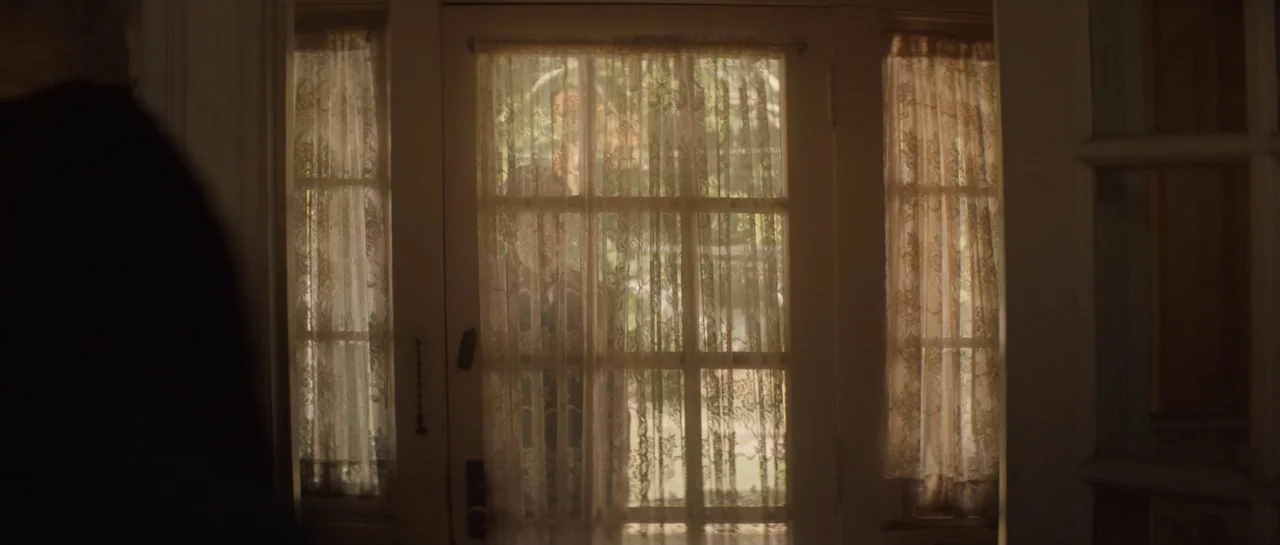 The Front Room (2024) movie still: medium — A residential front door with glass panels covered by ornate lace curtains. A person is visible through…