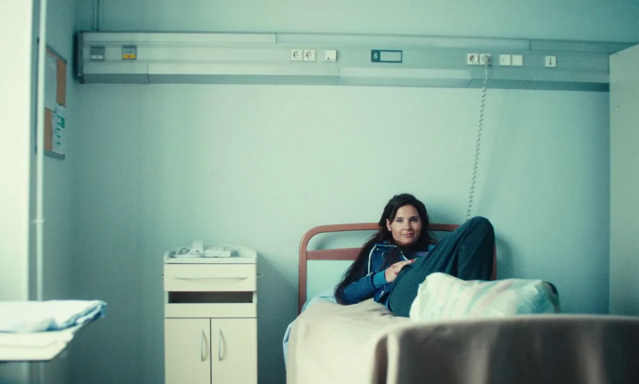 Just The Two Of Us (2023) movie still: medium — A sterile medical room features a light blue wall with a mounted service panel. A woman lies in a…