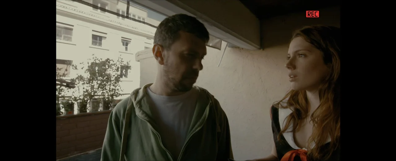 Faraday (2013) movie still: medium — A man and a woman stand on a building balcony during the daylight. The pair are engaged in a conversation with…