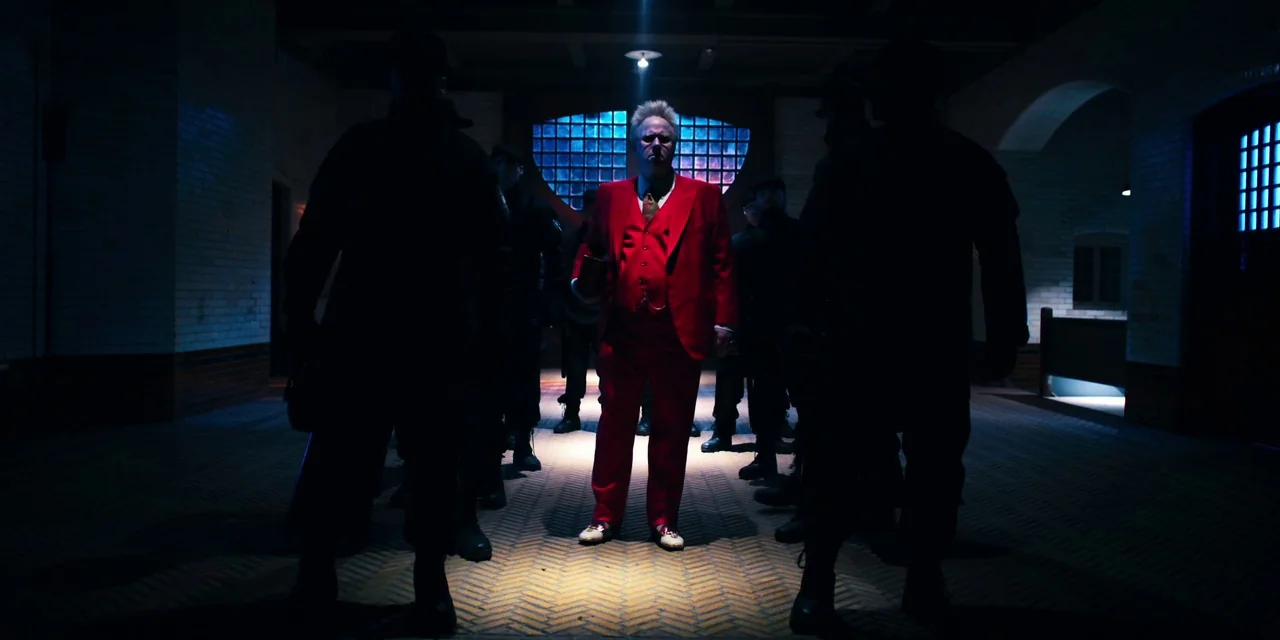 Polar (2019) movie still: fullbody — A dark, brick-walled hallway is dimly lit by blue ambient light. A man in a bright red suit stands in a central…