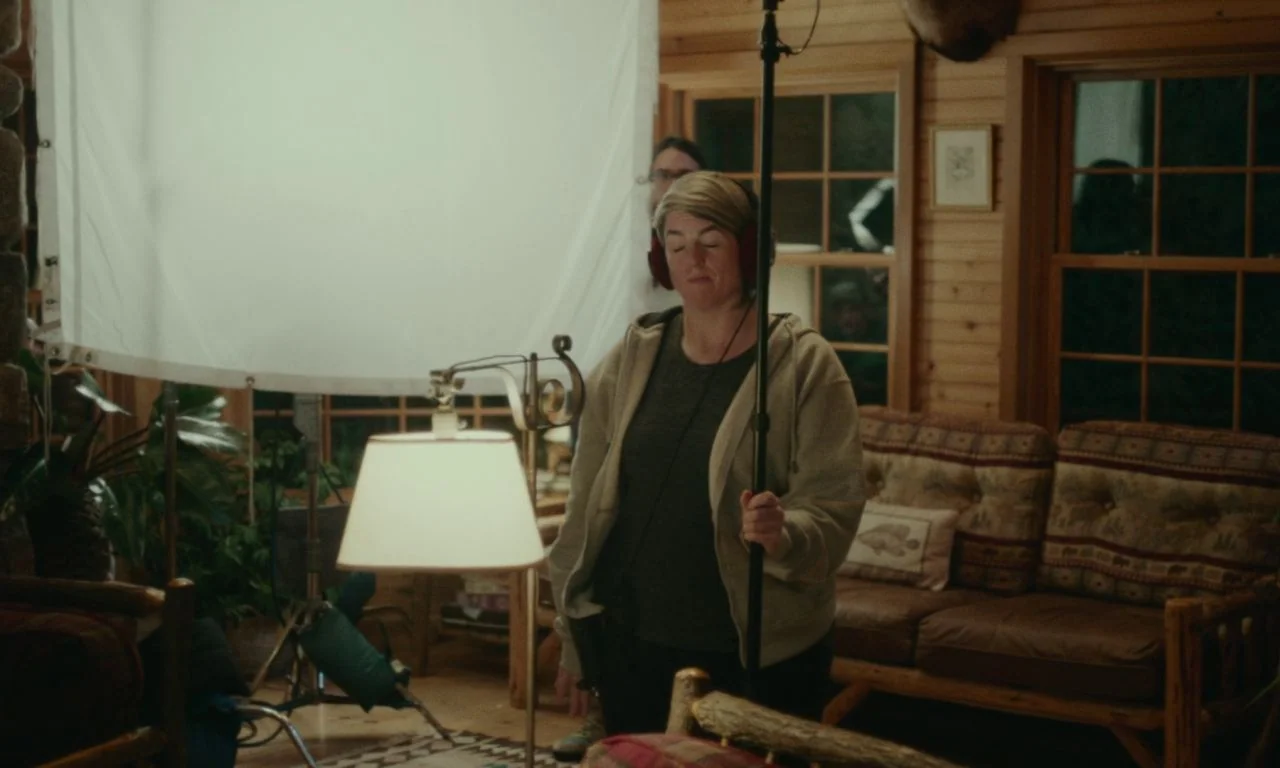 Black Bear (2020) movie still: medium — A rustic log cabin interior features wooden walls and a patterned couch at night. A sound technician stands…