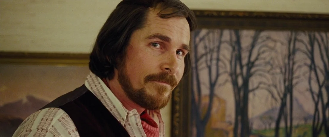 American Hustle (2013) movie still: closeup — An interior space displays framed landscape paintings against a warm-toned wall. A man with a dark…