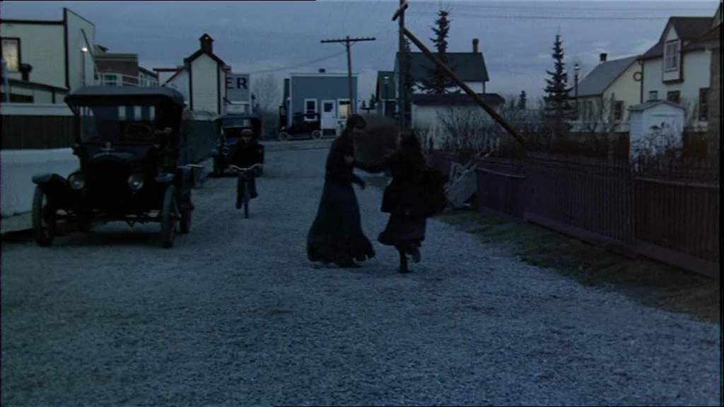 Days Of Heaven (1978) movie still: wide — A gravel road runs between wooden houses under a dim, overcast twilight sky. Two women rotate while…