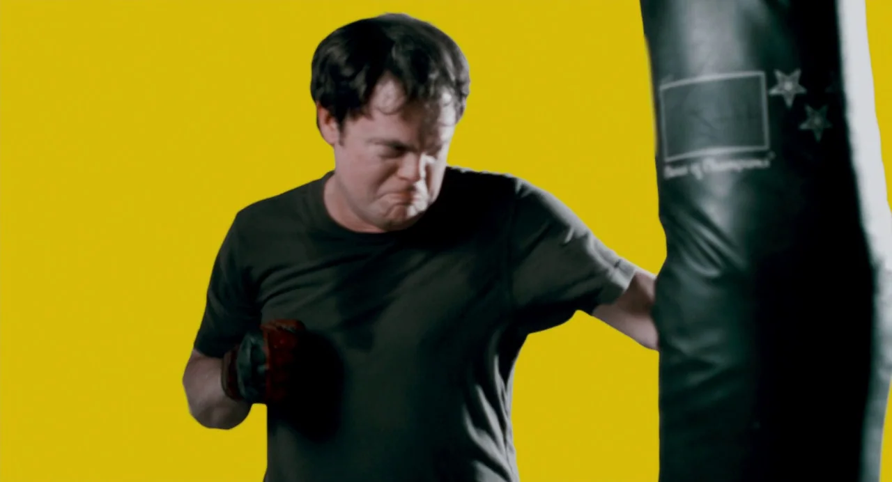 Super (2010) movie still: medium — A man stands in front of a bright solid yellow studio backdrop. He wears gloves and hits a black hanging heavy bag.