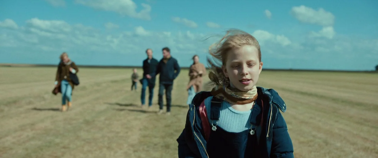 Speak No Evil (2022) movie still: medium — A group of people walks through a flat, harvested agricultural field under a blue sky. A young girl leads…