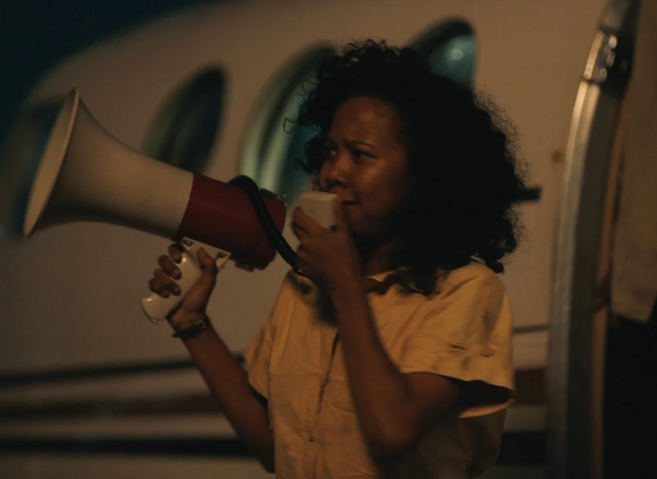 Red Island (2023) movie still: medium — An outdoor tarmac area at night with the side of an aircraft fuselage visible. A woman with curly hair holds…
