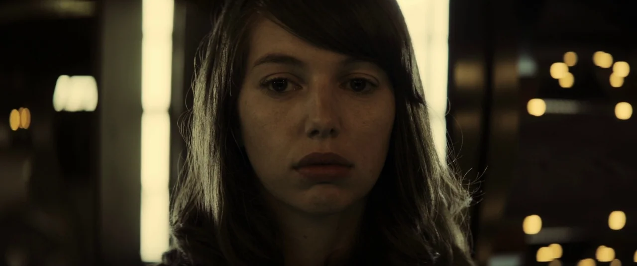 The Hole in the Ground (2019) movie still: closeup — A dimly lit indoor space features glowing rectangular columns flanking the subject. The woman…
