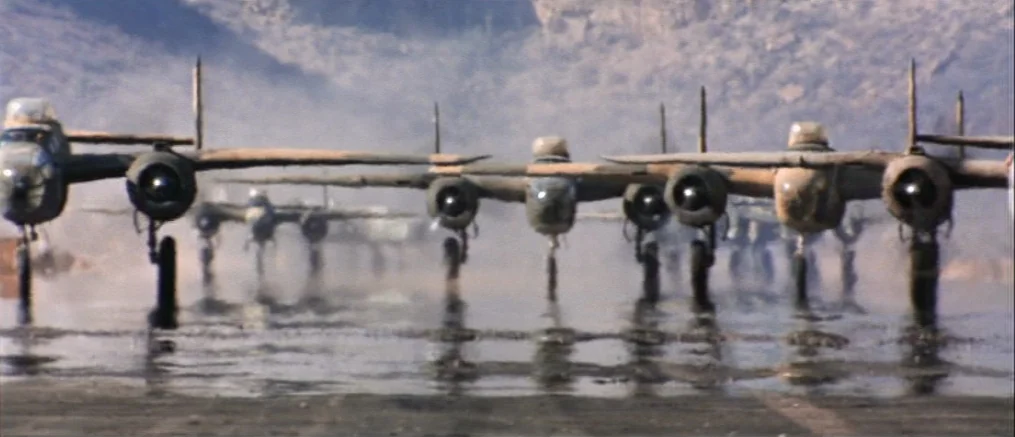 Catch 22 (1970) movie still: wide — A line of B-25 Mitchell bombers sits on a damp, reflective concrete runway. Dust and light haze obscure the…