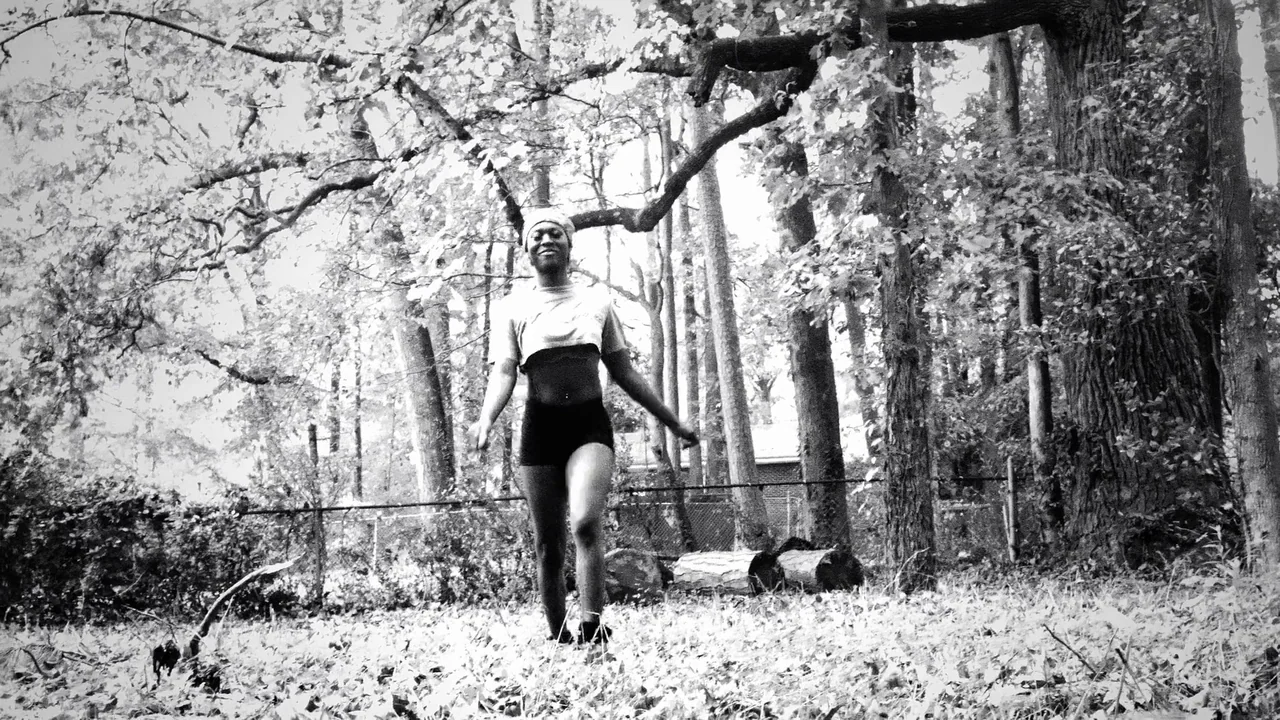 Kokomo City (2023) movie still: fullbody — A wooded area with mature trees and a chain-link fence visible in the background. A woman walks forward…