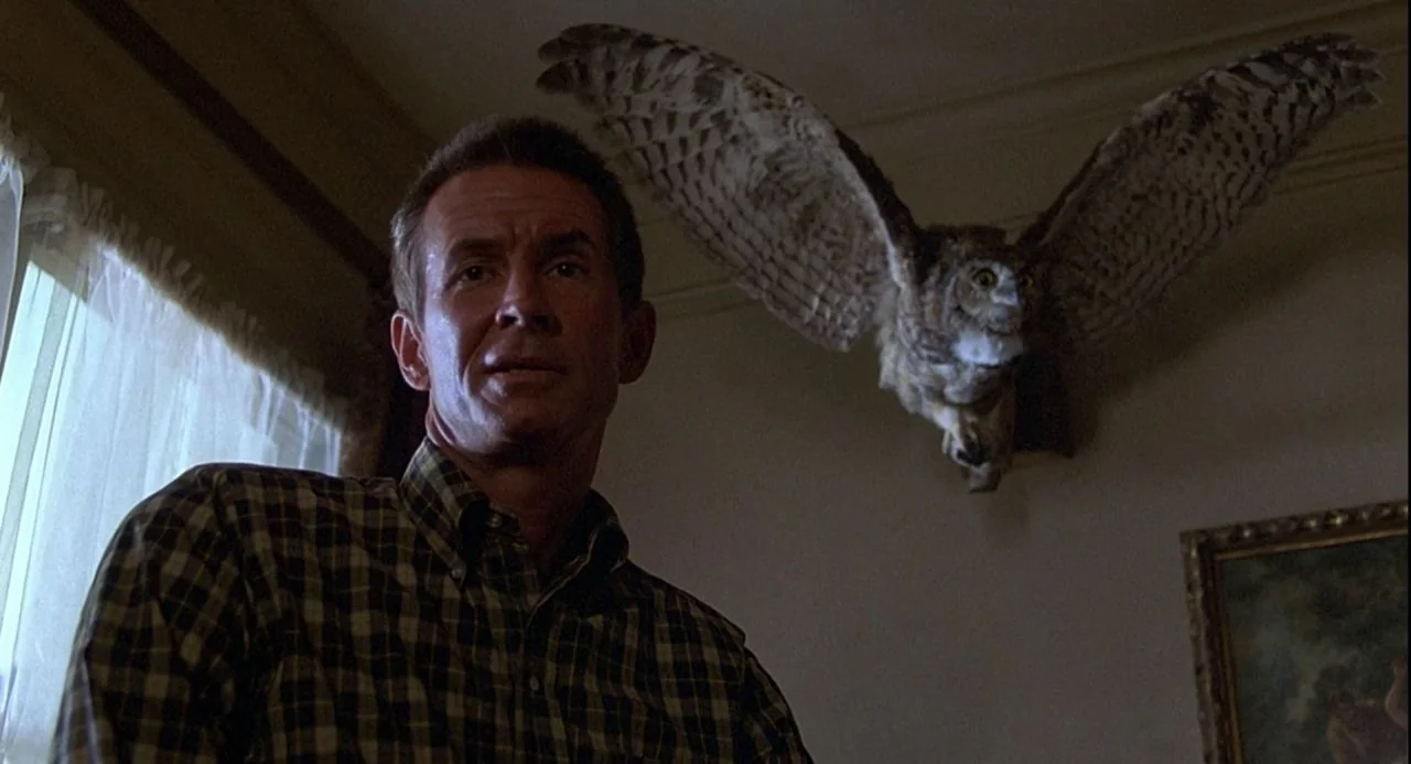 Psycho III (1986) movie still: medium — A dimly lit room features a taxidermied owl mounted high on a wall. A man in a plaid shirt stands in the…