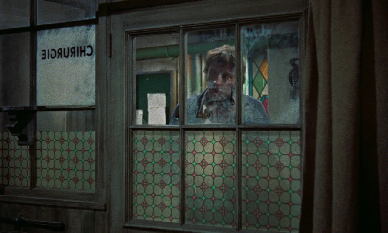 The Revenge of Frankenstein movie still: medium — A small clinical interior space features a wooden door with decorative glass panes. A man stands…