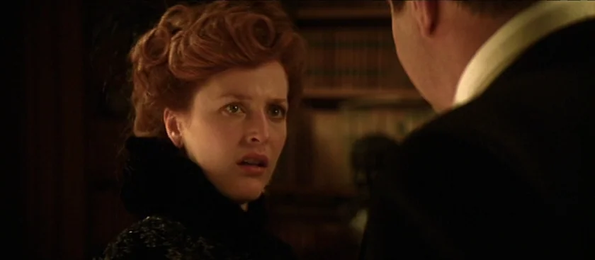 The House of Mirth (2000) movie still: medium — A dimly lit room with bookshelves visible in the blurred background. A red-haired woman gazes toward…