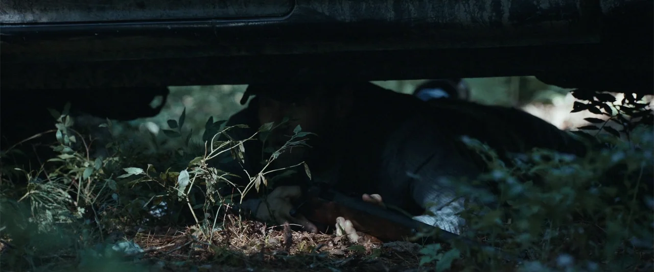 It Comes at Night (2017) movie still: medium — A man lies prone under the chassis of a vehicle in a wooded area. He holds a rifle and looks ahead…