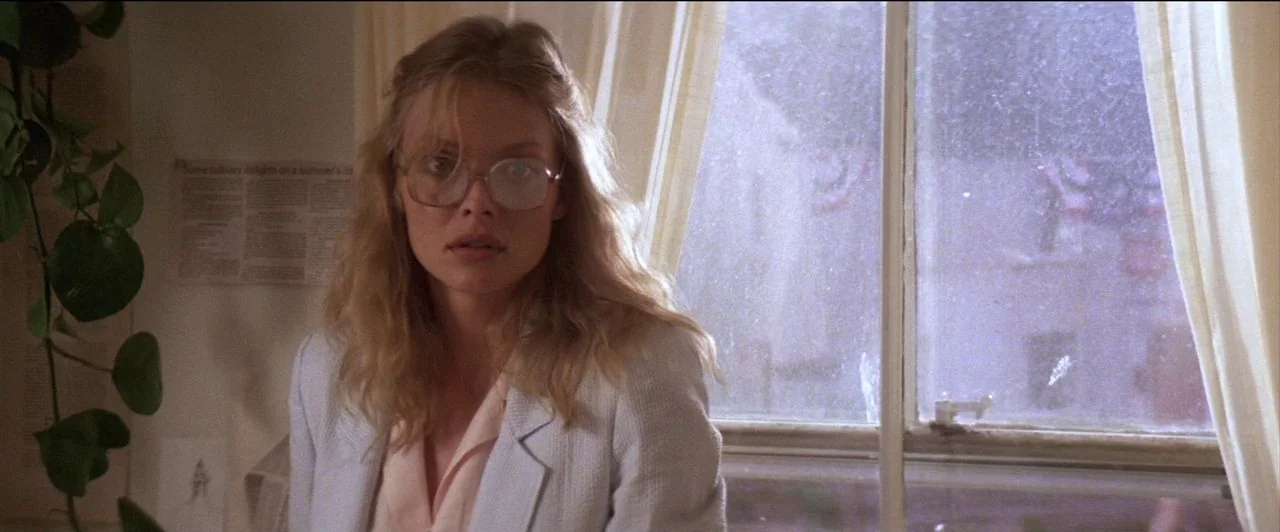 The Witches of Eastwick (1987) movie still: medium — A sunlit room features a window with sheer curtains and a trailing houseplant. A woman with…