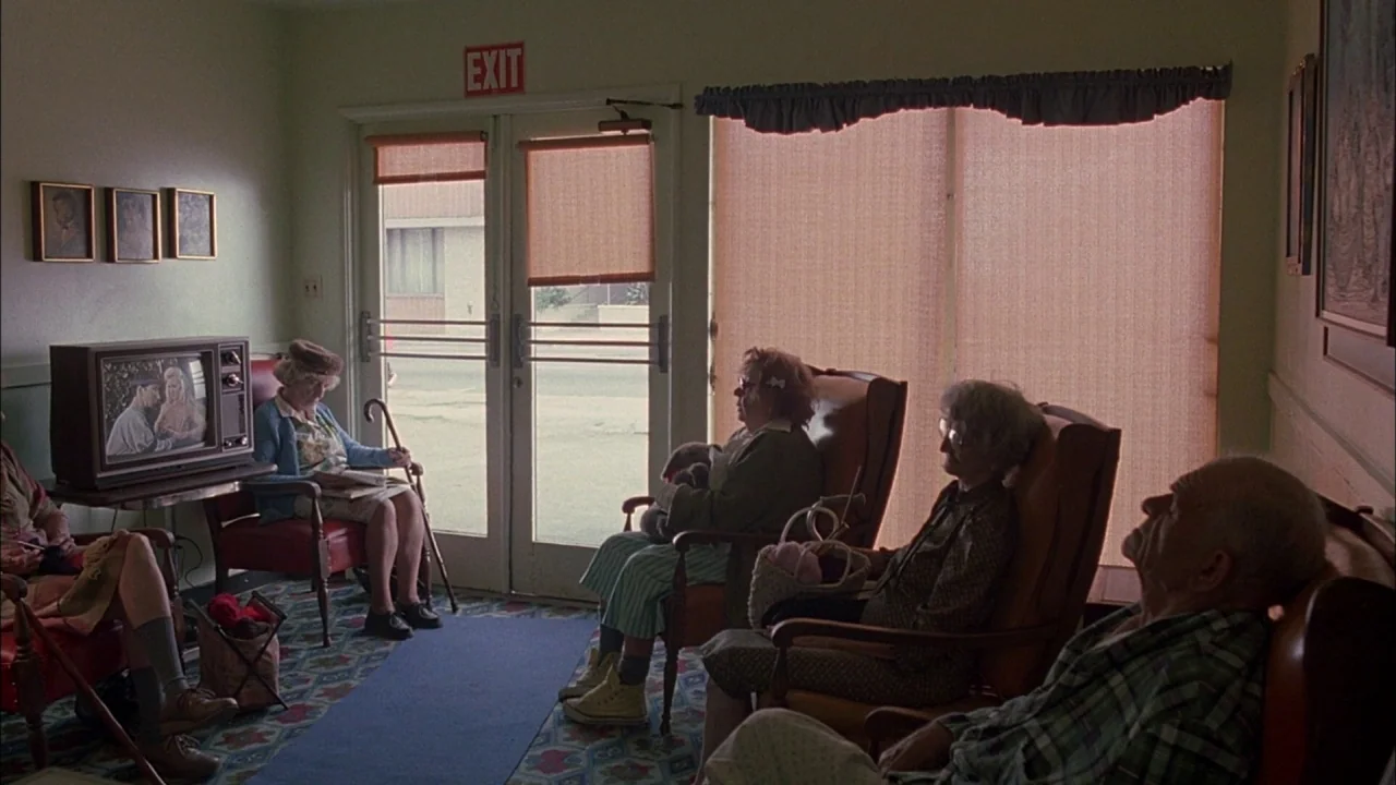 Whore (1991) movie still: wide — A nursing home lounge with patterned carpeting and armchairs arranged before a television. Elderly residents sit in…