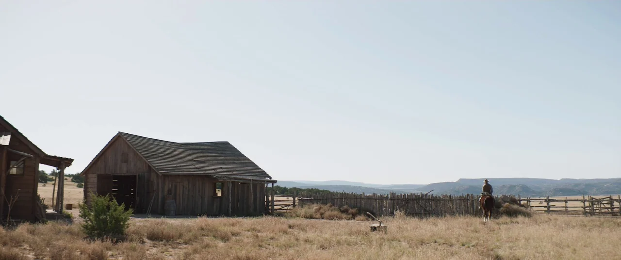 The Wind (2018) movie still: wide — A rural western homestead features weathered wooden buildings and a rustic stick fence. A single horseman rides…