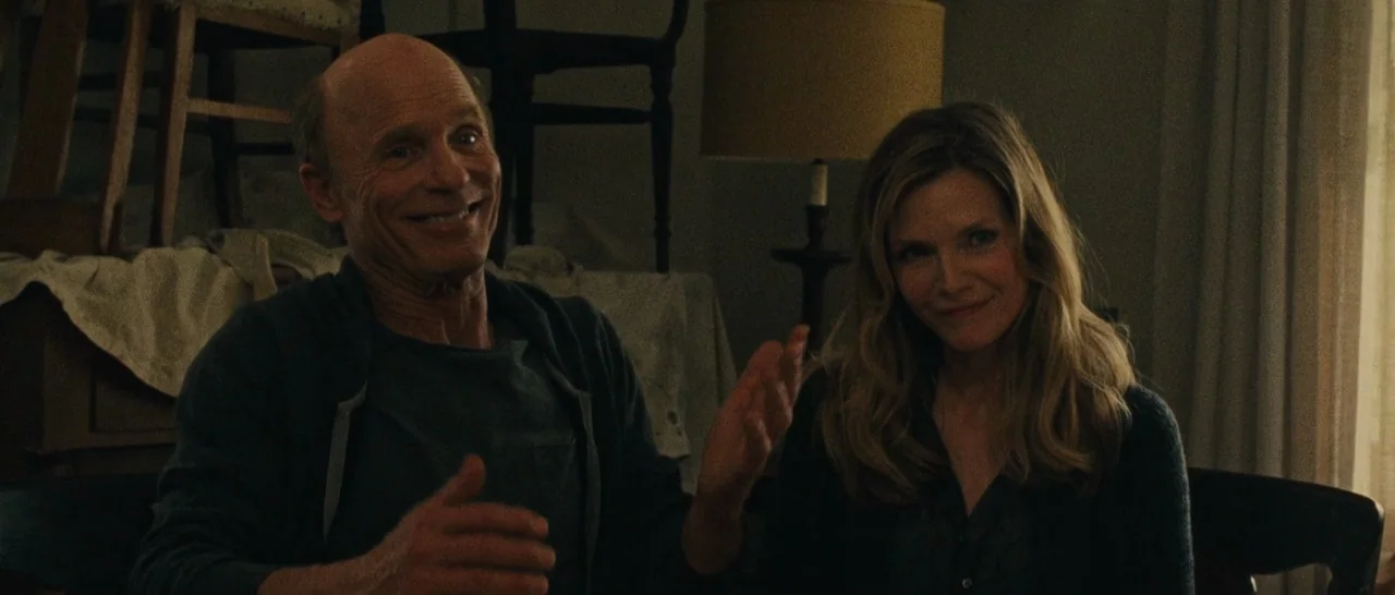 mother! (2017) movie still: medium — A dimly lit room contains wooden chairs stacked with draped white cloths in the background. An older man and a…