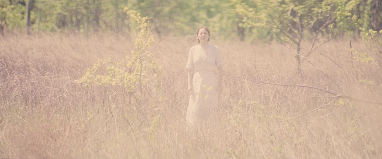 The Immigrant (2013) movie still: fullbody — A woman stands in the middle of a vast, tall-grass field during daylight. Young trees are scattered…