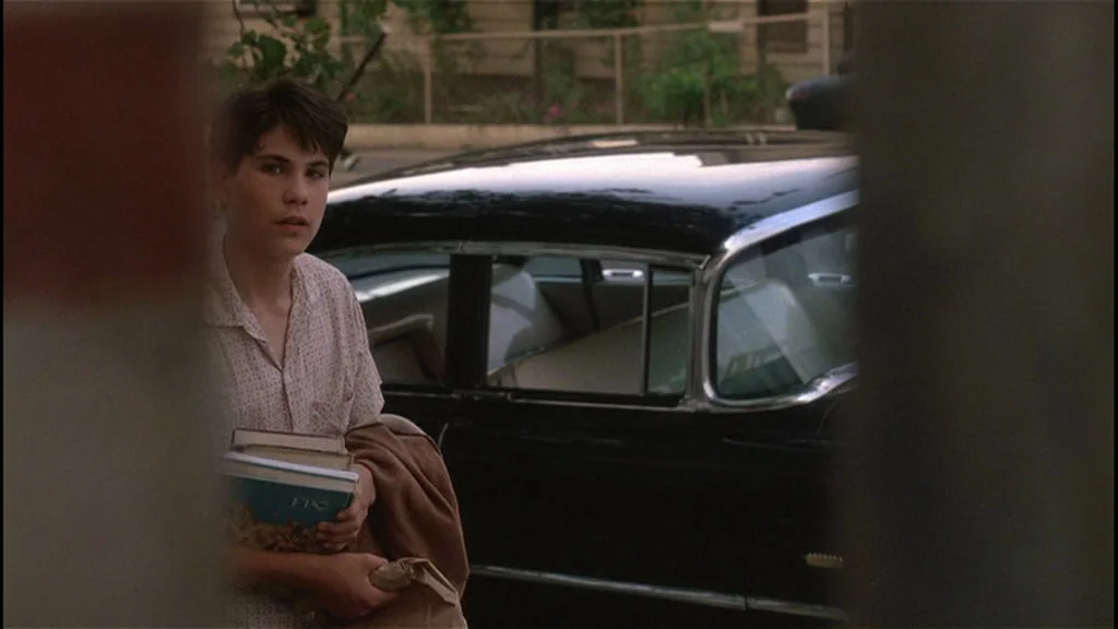 Goodfellas (1990) movie still: medium — A teenage boy stands next to a vintage black car on a suburban street. He holds a stack of books and a brown…