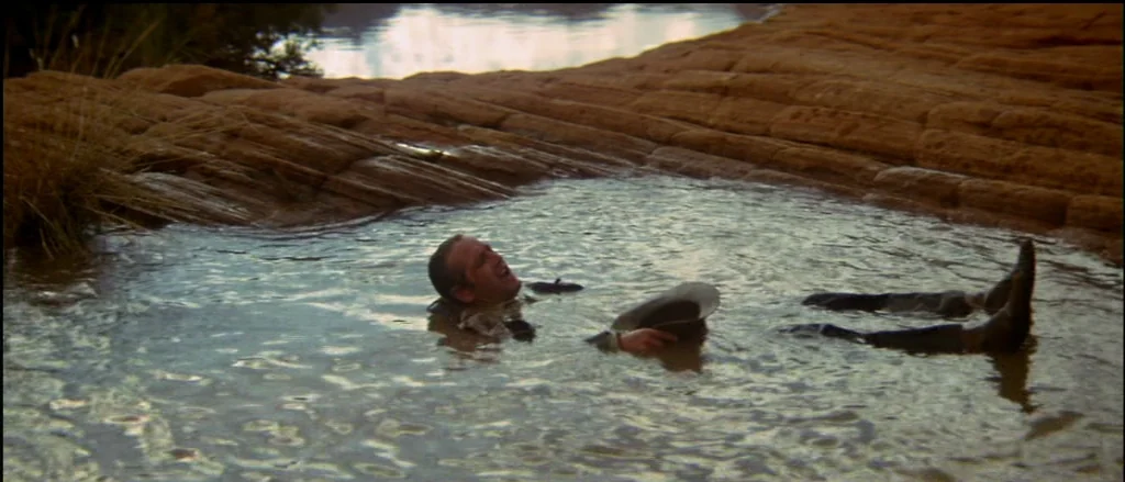 Butch Cassidy and the Sundance Kid (1969) movie still: wide — A man lies partially submerged in a small, murky pool surrounded by textured sandstone…