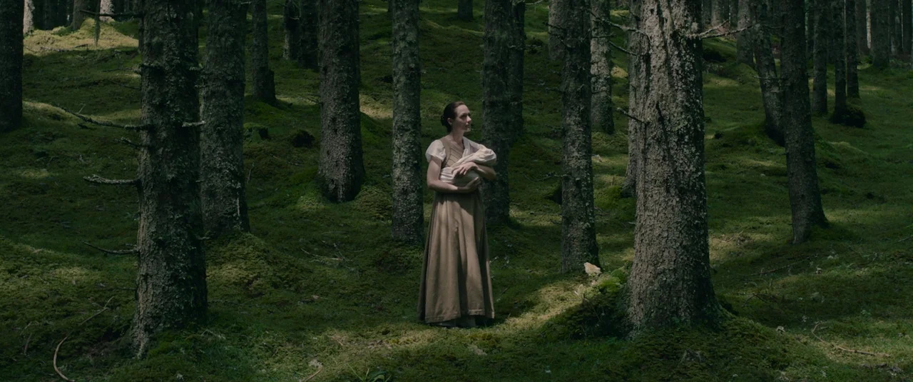 Hagazussa (2017) movie still: fullbody — A dense coniferous forest floor covered in thick green moss with numerous tall trees. A woman in a simple…