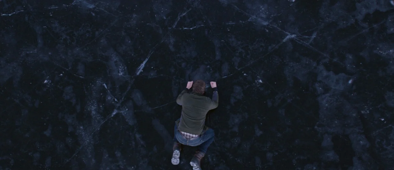 Thelma (2017) movie still: wide — A vast expanse of thick, cracked dark ice covers a frozen body of water. A man kneels in the center of the frame,…