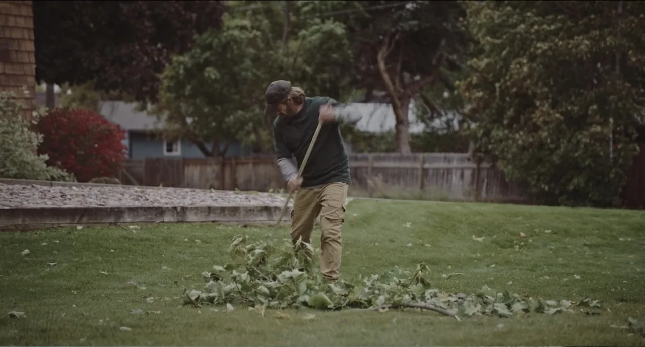 Cowboys (2014) movie still: fullbody — A residential backyard features a mowed green lawn surrounded by mature leafy trees. A man in a dark shirt…